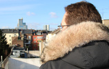 Ein Mann im Mantel mit Pelzkragen an der Kapuze blickt von einem Balkon auf Häuser. Bild: Verbraucherzentrale NRW