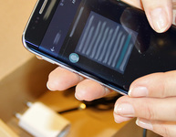 Zwei Hände umfassen ein Smartphone über einem Karton mit Zubehör. Foto: hamo