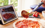 Tomatensoße wird mit einer Kelle auf einer Pizza verteilt. Dahinter steht ein Tablet, auf dem eine Rezepte-App läuft. Bild: Monkey Business / Fotolia.com