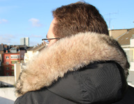 Ein Mann im Mantel mit Pelzkragen an der Kapuze blickt von einem Balkon auf Häuser. Bild: Verbraucherzentrale NRW