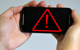 Ein rotes Ausrufezeichen im Dreieck auf dem Bildschirm eines Smartphones, gehalten von zwei Händen. Bild: checked4you
