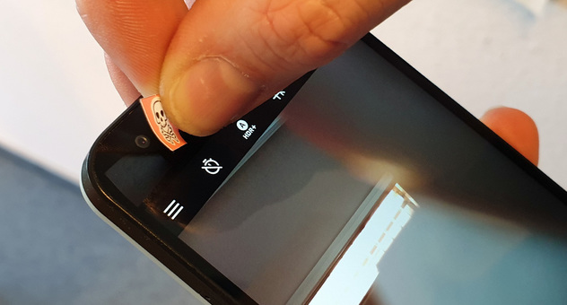 Zeigefinger und Daumen kleben einen kleinen Aufkleber mit Totenkopf auf die Selfie-Cam eines Smartphones. Bild: Verbraucherzentrale NRW