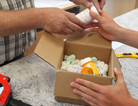 Hände nehmen eine Pillendose aus einem Paket. Foto: Zollarchiv/Verbraucherzentrale NRW